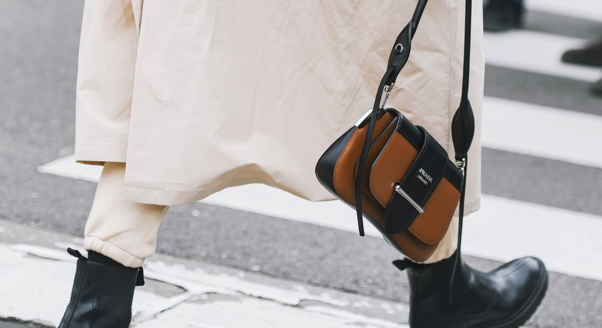 Schwarze klobige Stiefel und beige Hose unter langem Mantel, Person hält eine braun-schwarze Designerhandtasche, Gehweg im Hintergrund.