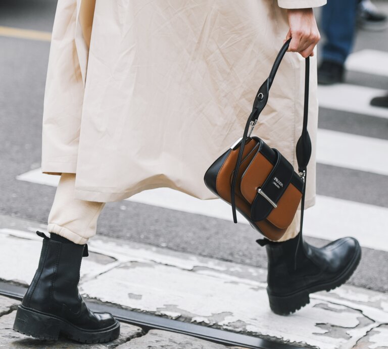 Schwarze klobige Stiefel und beige Hose unter langem Mantel, Person hält eine braun-schwarze Designerhandtasche, Gehweg im Hintergrund. | © Adobe Stock/ agcreativelab
