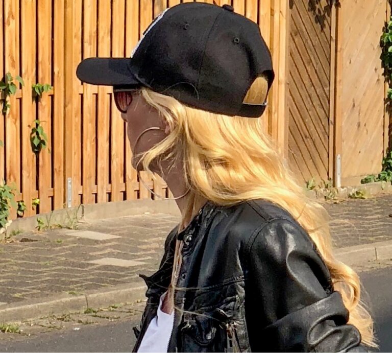 Seitenansicht einer Person mit lässigem Look aus Lederjacke, Cap und Sonnenbrille, umgeben von sommerlicher Szenerie an einem Holzzaun. | © Renate Zott