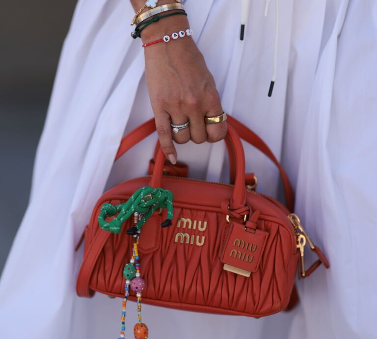 Hand mit goldenen und bunten Armbändern hält eine kleine orangene Miu Miu-Tasche mit dekorativen Elementen, vor einem weißen Kleid. | © Jeremy Moeller/Getty Images