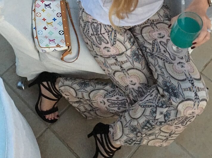 Eine Frau in bedruckter Hose, schwarzen High Heels und weißem Shirt hält ein türkisfarbenes Getränk, daneben eine Designer-Handtasche. | © Renate Zott