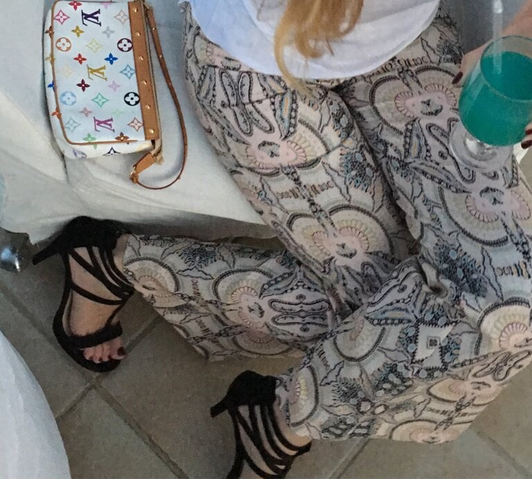 Eine Frau in bedruckter Hose, schwarzen High Heels und weißem Shirt hält ein türkisfarbenes Getränk, daneben eine Designer-Handtasche. | © Renate Zott