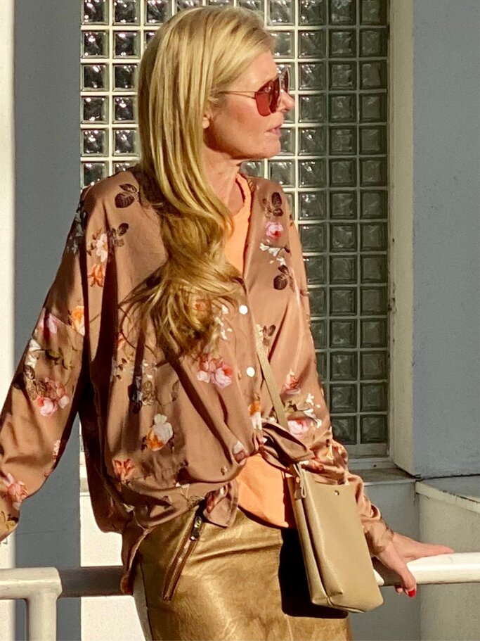 Eine Frau mit langen blonden Haaren trägt eine rosafarbene Bluse mit Blumenmuster, einen goldenen Rock und eine kleine beigefarbene Tasche. | © Renate Zott