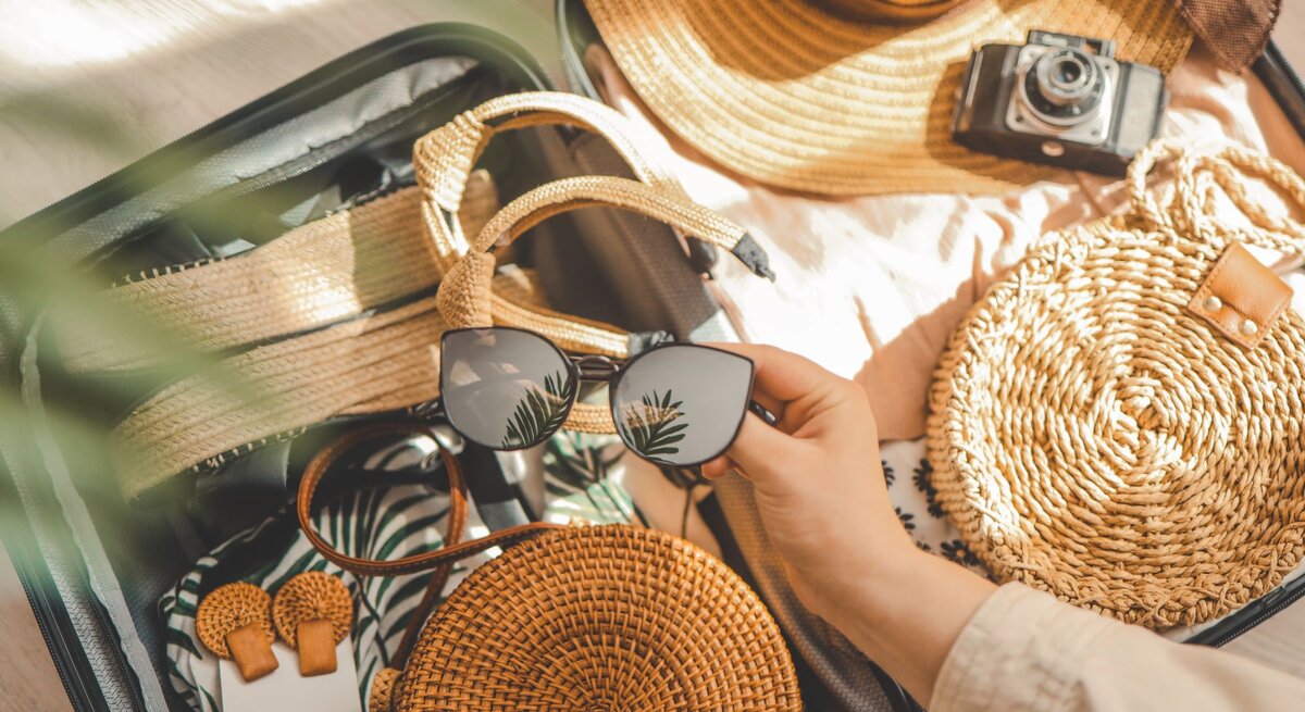 Offener Reisekoffer gefüllt mit Strandkleidung, Sommer-Sandalen, runder Basttasche, Sonnenhut und Hand mit schwarzer Sonnenbrille mit Pflanzenreflex.