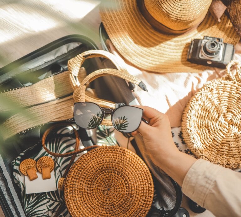 Offener Reisekoffer gefüllt mit Strandkleidung, Sommer-Sandalen, runder Basttasche, Sonnenhut und Hand mit schwarzer Sonnenbrille mit Pflanzenreflex. | © Adobe Stock/  shine.graphics