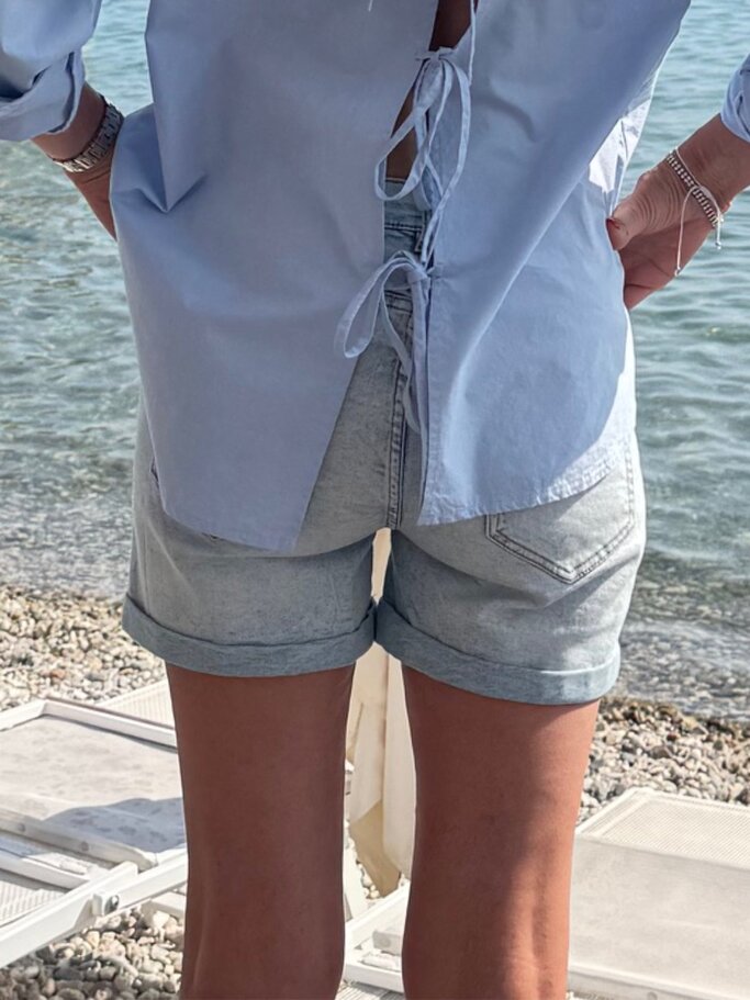 Detailaufnahme einer Frau in casual Shorts und leichter Bluse mit Rückenschleifen vor einem sonnigen Meereshintergrund. | © Renate Zott
