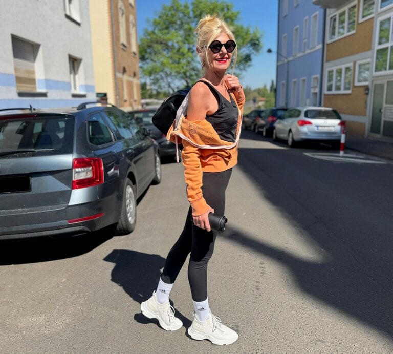 Renate Zott mit Sonnenbrille, orangefarbener Jacke und schwarzem Sportoutfit läuft lachend mit Rucksack durch Wohnstraße. | © Renate Zott 