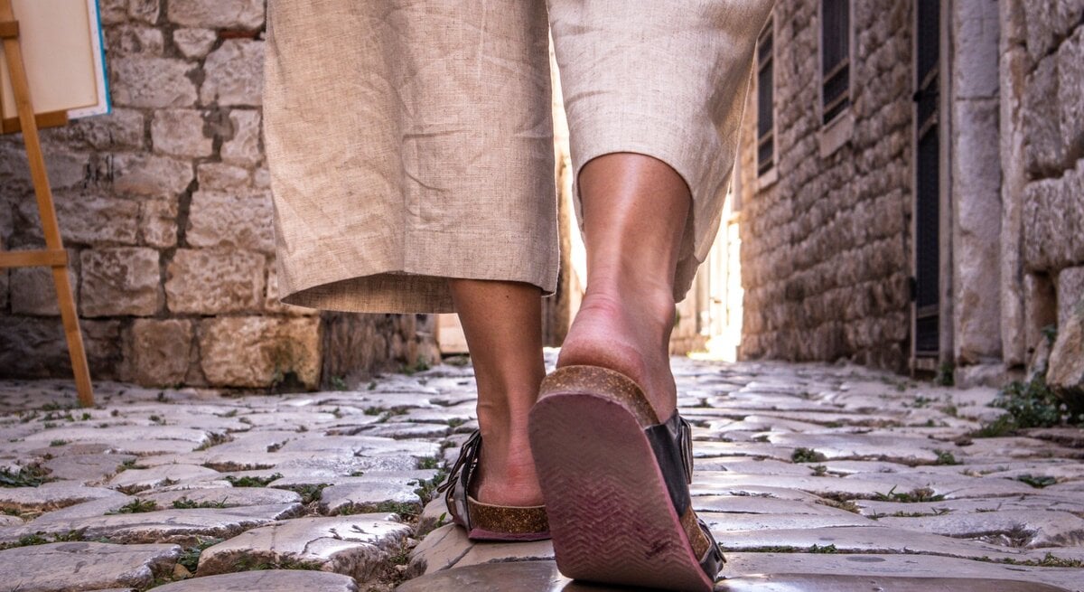 Nahaufnahme von Beinen in beigen Leinenhosen und Sandalen, die über glänzende Kopfsteinpflaster in einer engen Altstadtgasse laufen.