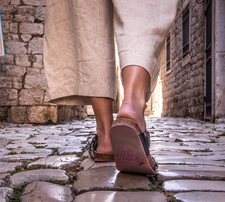 Nahaufnahme von Beinen in beigen Leinenhosen und Sandalen, die über glänzende Kopfsteinpflaster in einer engen Altstadtgasse laufen. | © Adobe Stock/ kasto