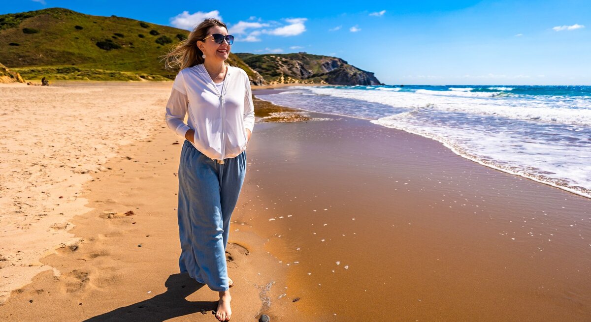 Eine Frau in weißer Bluse und blauer Hose spaziert barfuß am sonnigen Sandstrand entlang, während kleine Wellen ans Ufer schlagen.