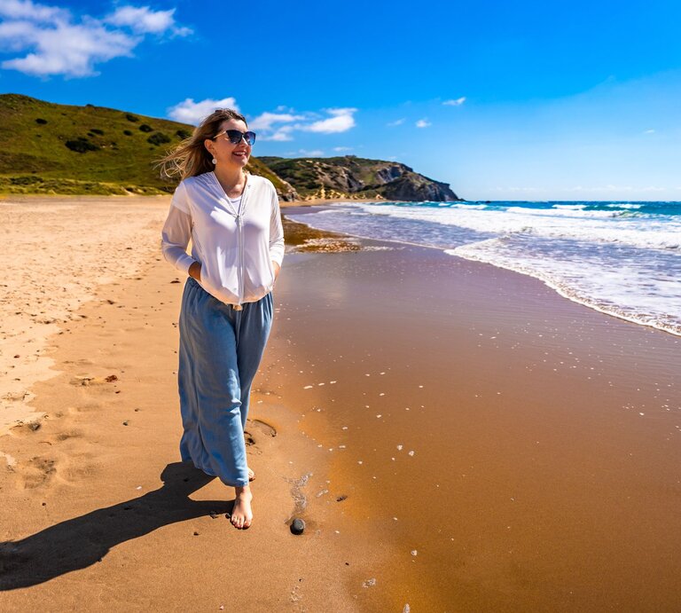 Eine Frau in weißer Bluse und blauer Hose spaziert barfuß am sonnigen Sandstrand entlang, während kleine Wellen ans Ufer schlagen. | © Adobe Stock/ Jacek Chabraszewski