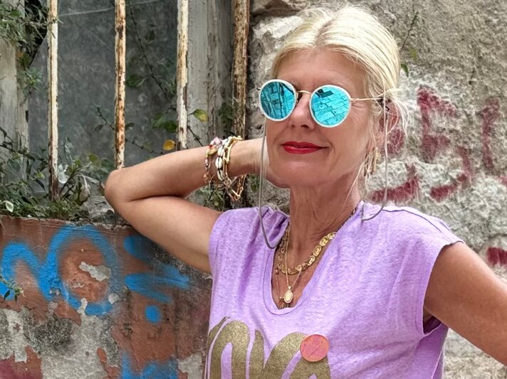 Renate Zott mit türkis verspiegelter Sonnenbrille, lilafarbenem Shirt und goldenen Ketten steht entspannt vor Graffitiwand. | © Renate Zott
