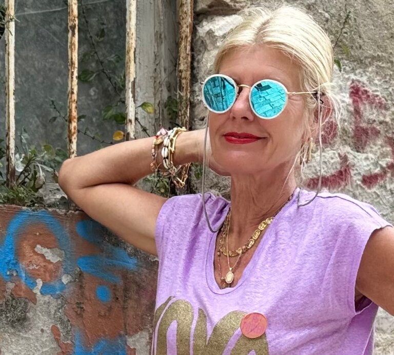 Renate Zott mit türkis verspiegelter Sonnenbrille, lilafarbenem Shirt und goldenen Ketten steht entspannt vor Graffitiwand. | © Renate Zott