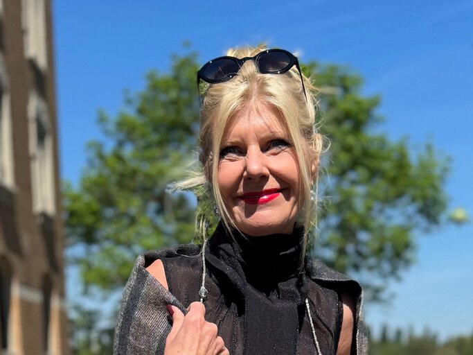 Anti-Aging-Accessoires: Renate Zott mit Hochsteckfrisur, rotem Lippenstift und schwarzer Sonnenbrille im Haar steht lächelnd vor blauem Himmel. | © Renate Zott