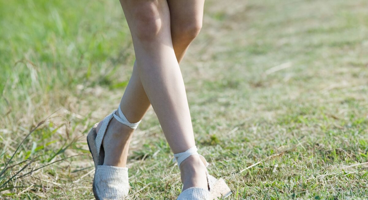 Junge Frau mit gebräunten Beinen geht barfuß in weißen Espadrilles-Sandalen über einen sonnigen Grasweg, grünes Sommerkleid weht leicht.