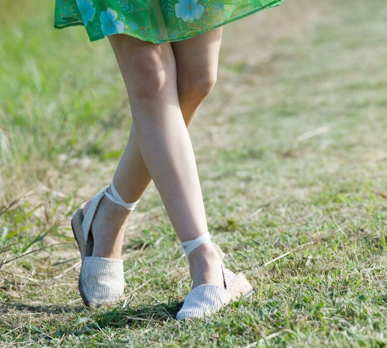 Junge Frau mit gebräunten Beinen geht barfuß in weißen Espadrilles-Sandalen über einen sonnigen Grasweg, grünes Sommerkleid weht leicht. | © Getty Images/ ZenShui/Laurence Mouton