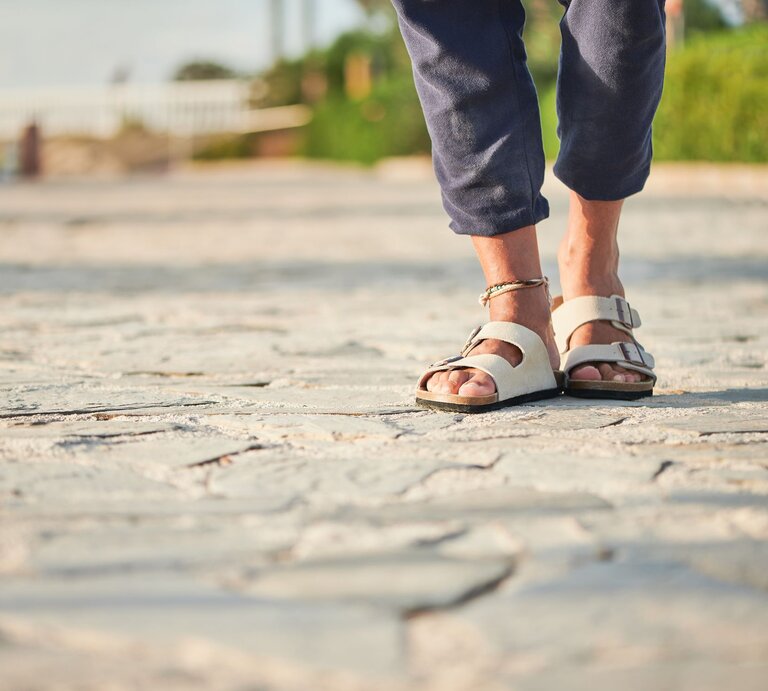 Füße in beigen Birkenstock-Sandalen und hochgekrempelter Hose auf heller Natursteinpromenade bei warmem Abendlicht. | © Adobe Stock/ Itxu