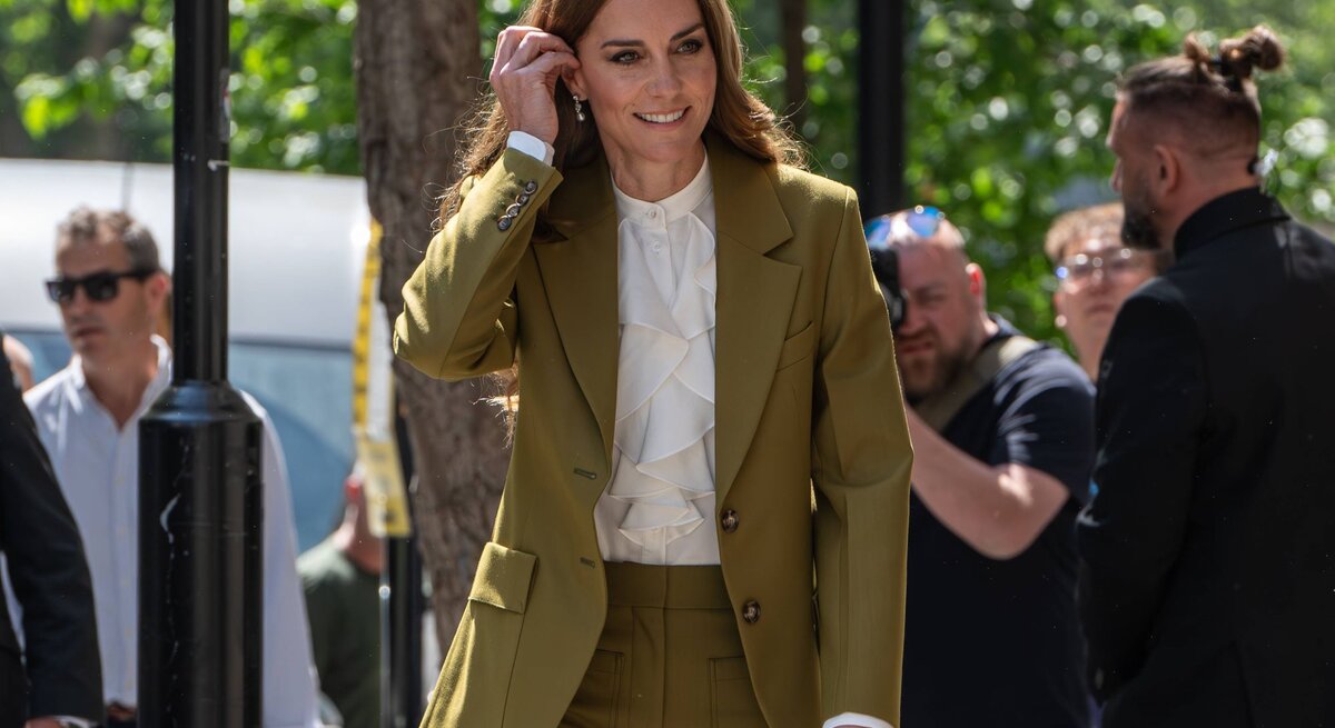 Prinzessin Kate  in olivgrünem Hosenanzug, Wedges Sandalen und weißer Rüschenbluse lächelt in die Kamera, umgeben von Fotografen und Passanten.