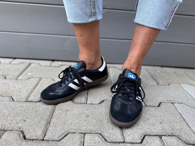 Schwarze Adidas-Sneaker mit weißen Streifen an gebräunten Füßen in gekrempelten Jeans auf grauem Pflasterstein. | © Renate Zott
