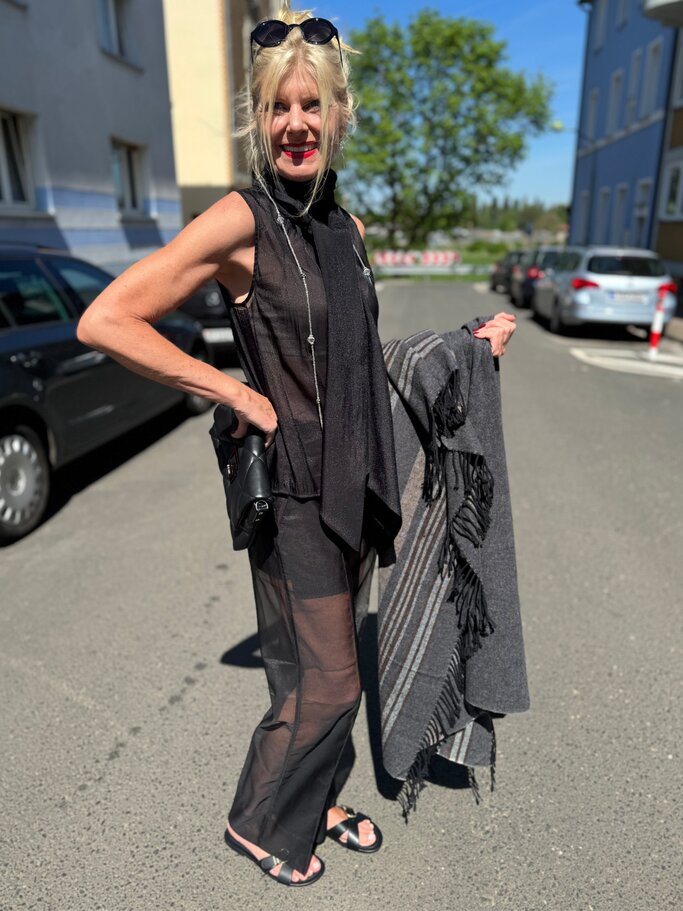 Renate Zott in schwarzem transparentem Ensemble mit Schal und Sandalen hält lächelnd einen grauen Fransenschal auf sonniger Straße. | © Renate Zott