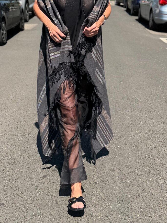 Schwarze transparente Hose unter grauem Fransencape mit Streifenmuster, kombiniert mit offenen Sandalen auf Asphalt bei Sonnenschein. | © Renate Zott