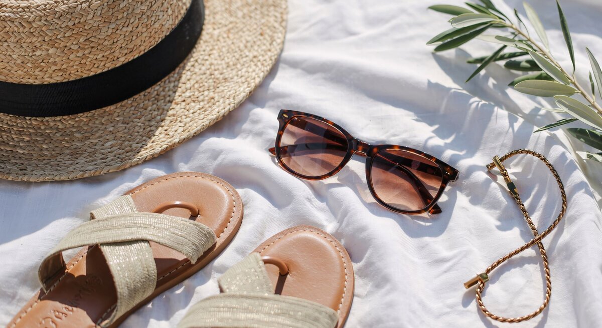 Strohhut, Sandalen, Sonnenbrille und ein goldenes Armband liegen sommerlich arrangiert auf weißem Tuch im Sonnenlicht.