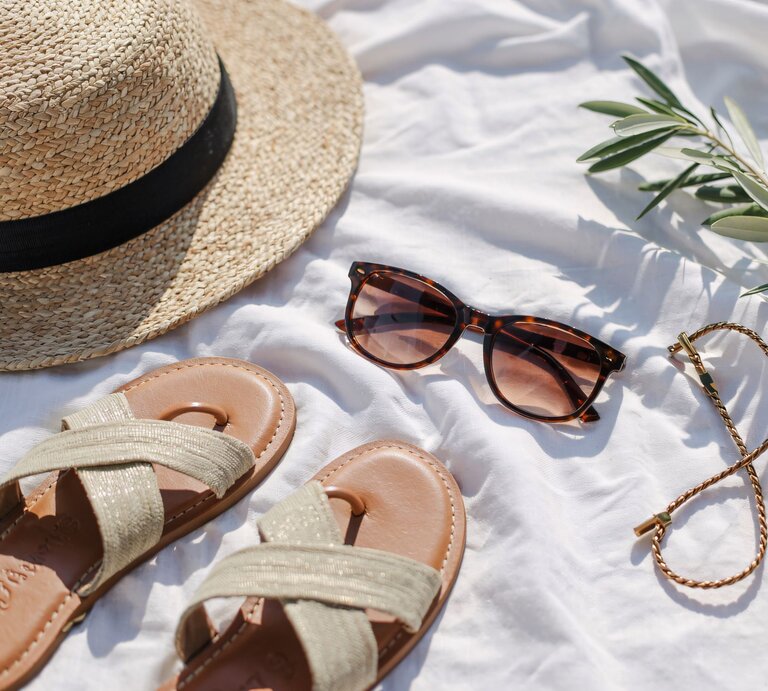 Strohhut, Sandalen, Sonnenbrille und ein goldenes Armband liegen sommerlich arrangiert auf weißem Tuch im Sonnenlicht. | © Adobe Stock/ StudioBackground