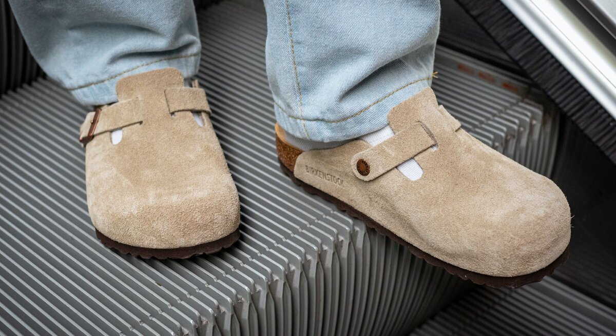 Zwei Füße in beigen Wildleder-Clogs und weißen Socken stehen auf einer fahrenden Rolltreppe aus Metallrillen.