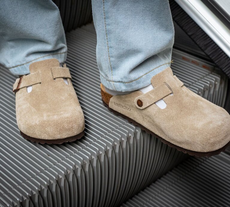 Zwei Füße in beigen Wildleder-Clogs und weißen Socken stehen auf einer fahrenden Rolltreppe aus Metallrillen. | © Adobe Stock/ David Bokuchava