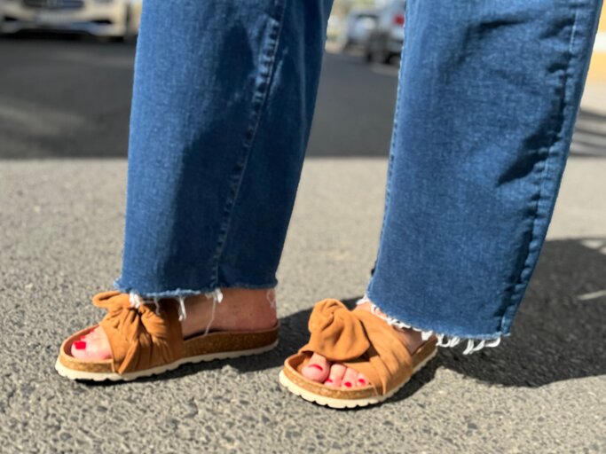 Füße einer Frau in braunen Sandalen mit Korksohle und rotem Nagellack auf Asphalt, Jeans mit ausgefranstem Saum. | © Renate Zott