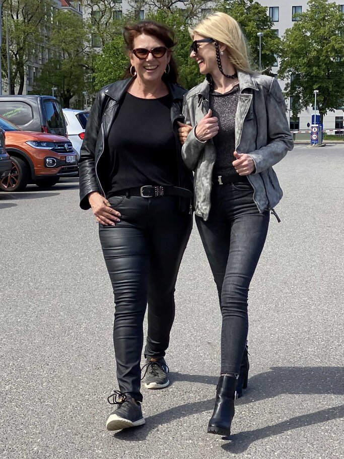 Zwei Frauen in Lederjacken und schwarzen Hosen gehen lachend Arm in Arm über einen Parkplatz an einem sonnigen Tag. | © Renate Zott