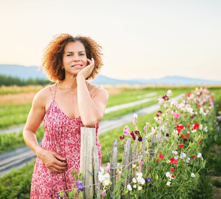 Eine fröhliche Frau in rotem Frühlingskleid steht zu Ostern an einem Blütenzaun auf dem Land, im Hintergrund Felder und sanfte Hügel. | © Shutterstock/ Anna Nahabed