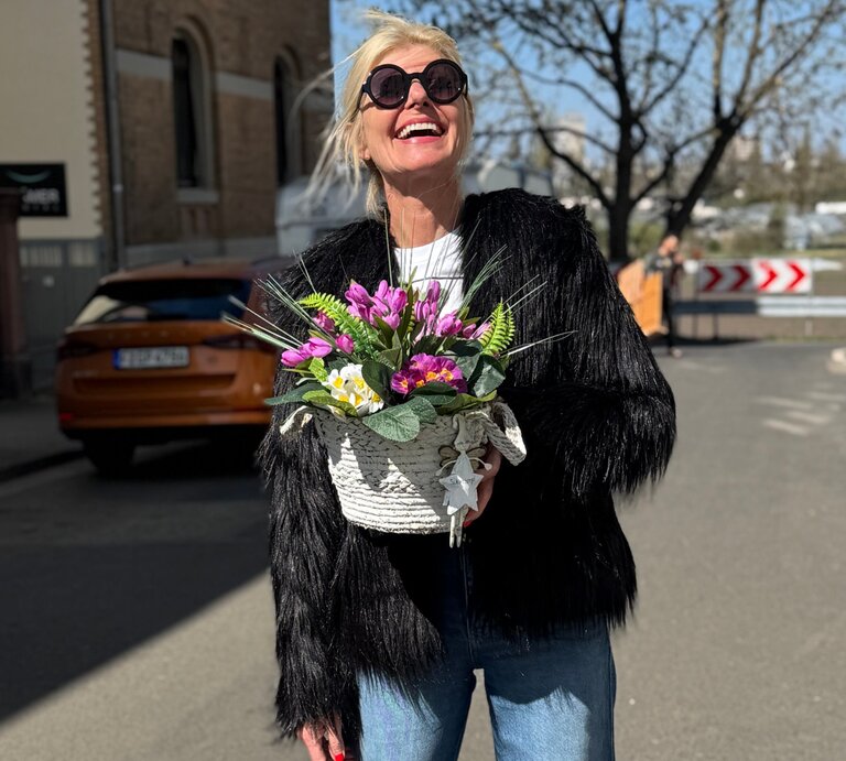 Lachende blonde Renate Zott mit schwarzer Fransjacke und Sonnenbrille hält Frühlingsblumenkorb auf sonniger Straße vor Stadtkulisse. | © Renate Zott 