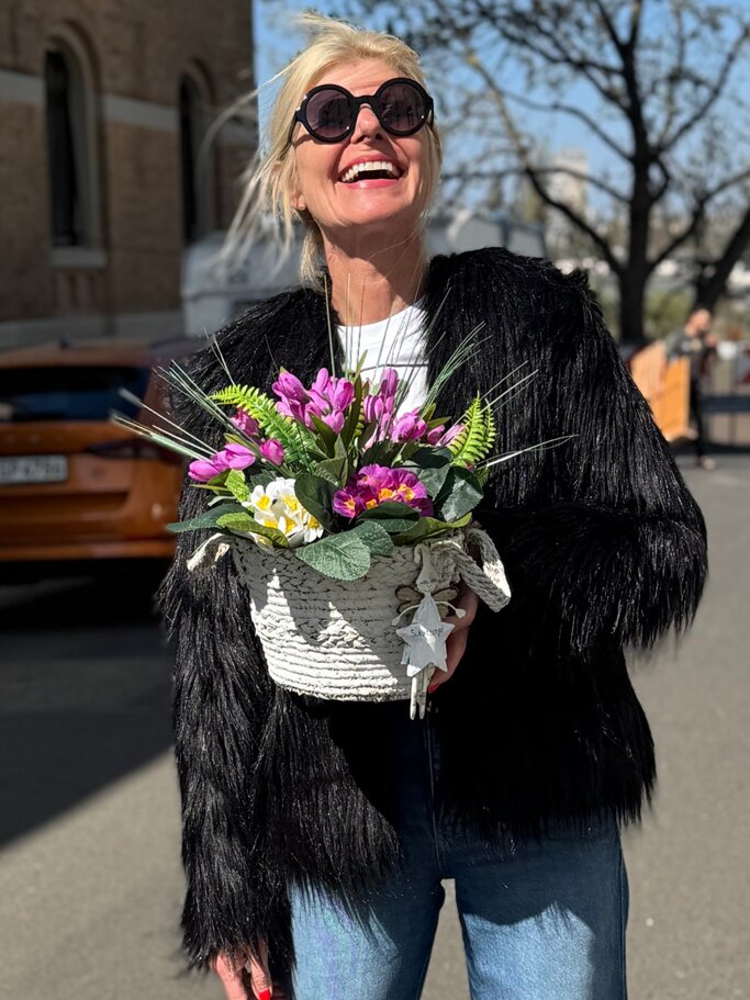 Lachende blonde Renate Zott mit schwarzer Fransjacke und Sonnenbrille hält Frühlingsblumenkorb auf sonniger Straße vor Stadtkulisse. | © Renate Zott 