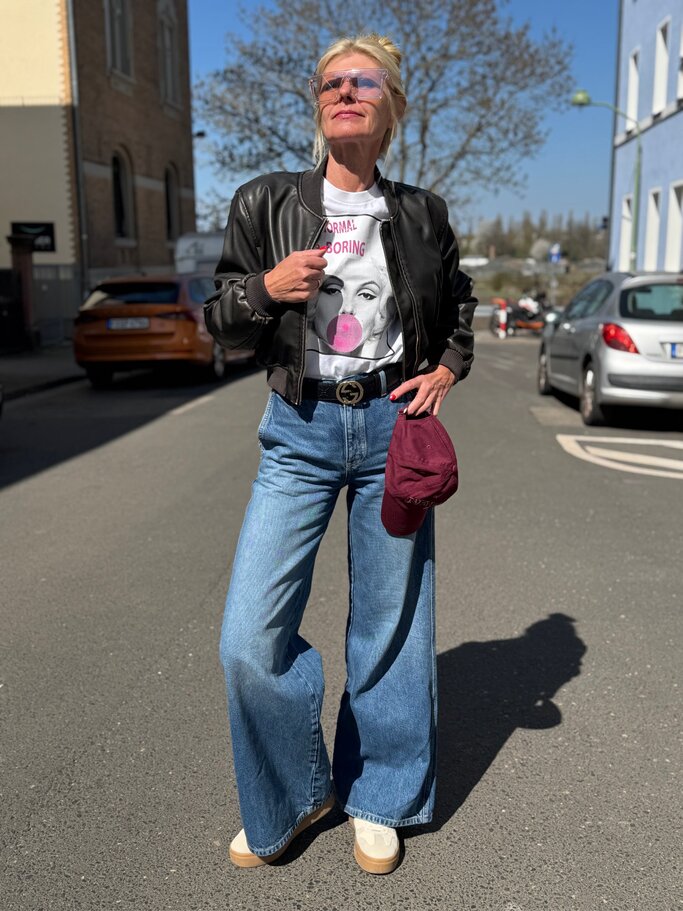 Blonde Renate Zott mit rosa Sonnenbrille trägt Leder-Bomberjacke, Pop-Art-Shirt und weite Jeans auf sonniger Straße. | © Renate Zott