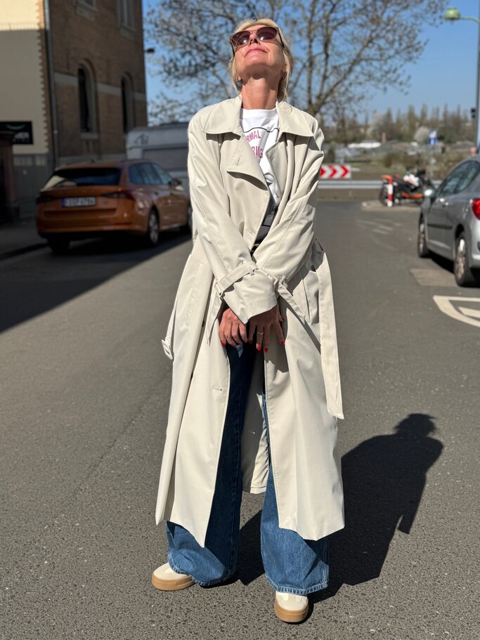 Blonde Renate Zott mit Sonnenbrille und langem beigem Trenchcoat blickt entspannt lächelnd in die Frühlingssonne auf leerer Straße. | © Renate Zott