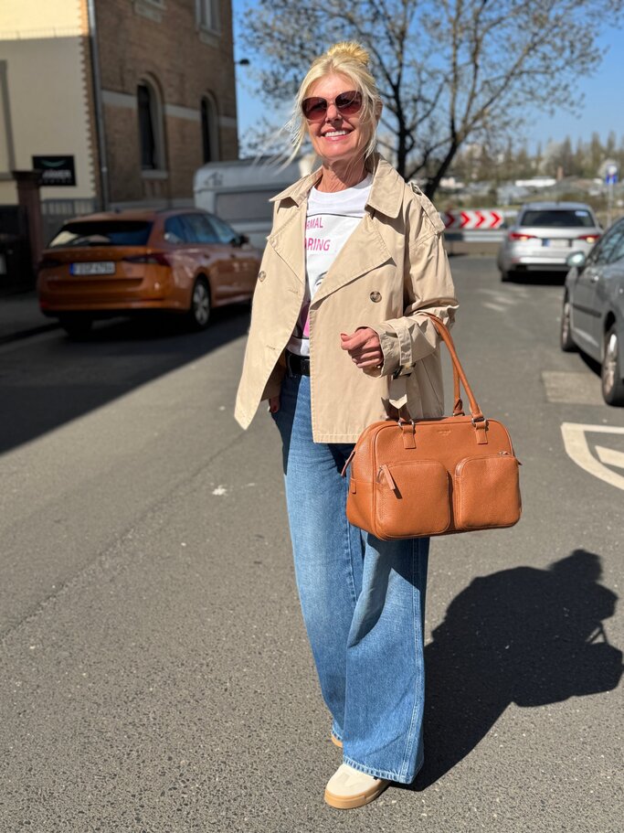 Lächelnde blonde Renate Zott mit Sonnenbrille trägt kurzen beigefarbenen Trenchcoat, weite Jeans und braune Lederhandtasche. | © Renate Zott