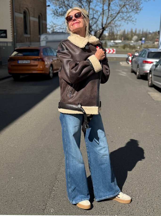 Blonde Renate Zott mit Sonnenbrille trägt braune Shearling-Jacke mit Teddykragen und weite Jeans auf sonniger Straße. | © Renate Zott