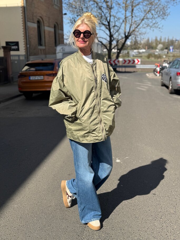Blonde Renate Zott mit Dutt trägt oversized olivgrüne Jacke, runde Sonnenbrille und weite Jeans auf sonniger Asphaltstraße. | © Renate Zott