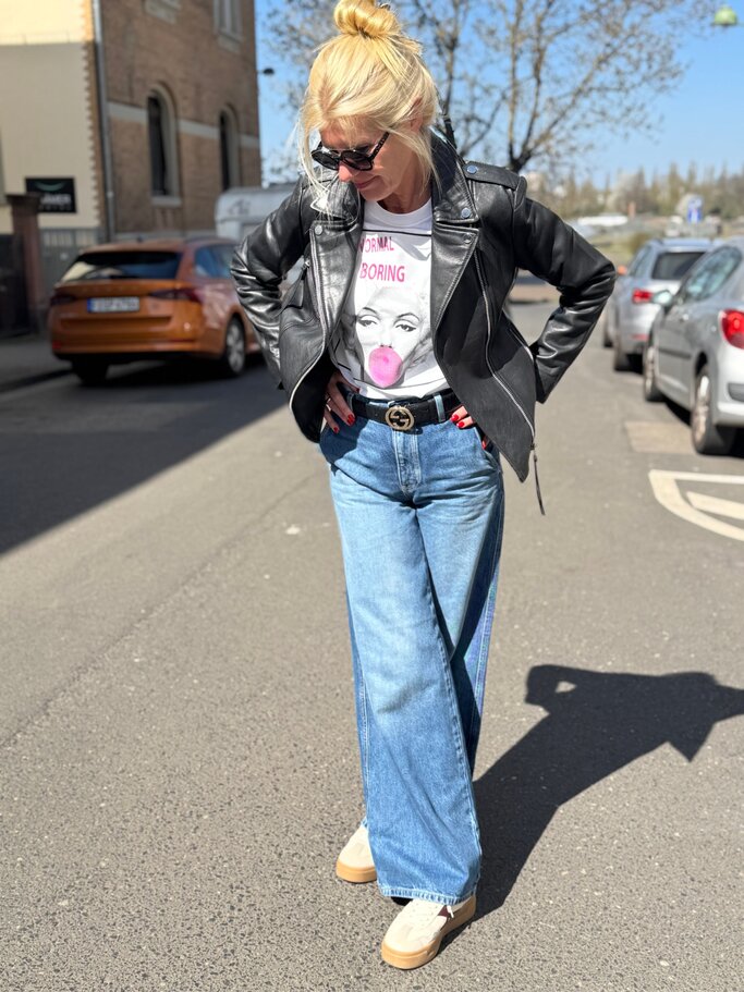 Blonde Renate Zott mit Dutt und Sonnenbrille trägt schwarze Lederjacke, Pop-Art-Shirt und weite Jeans auf sonniger Straße. | © Renate Zott