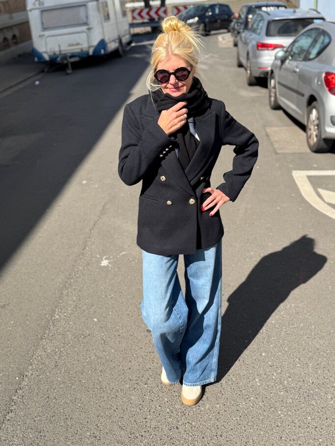 Blonde Renate Zott mit Dutt und schwarzer Sonnenbrille trägt dunklen Bouclé-Blazer, Schal und weite Jeans auf sonniger Straße. | © Renate Zott