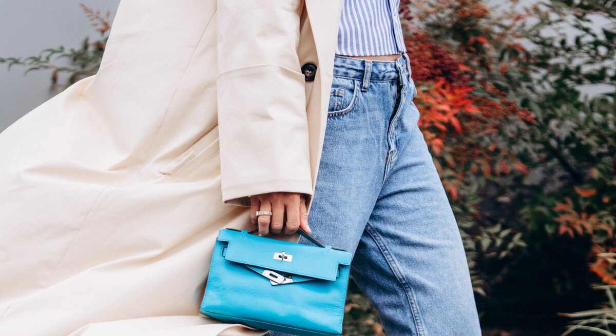 Zu sehen ist eine Frau, die einen Trenchcoat, einen kurzen dünnen Cardigan, eine Straight leg Jeans und eine hellblaue Tasche trägt.