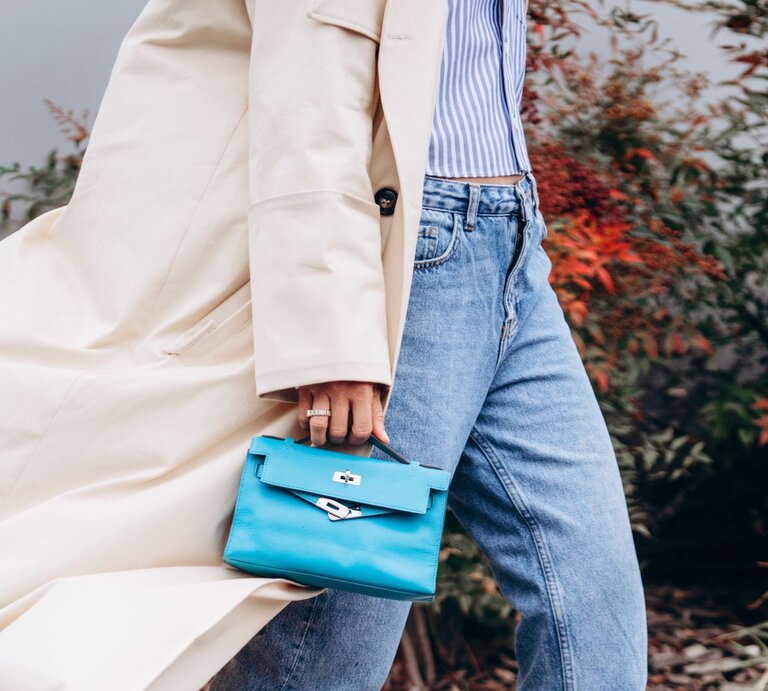 Zu sehen ist eine Frau, die einen Trenchcoat, einen kurzen dünnen Cardigan, eine Straight leg Jeans und eine hellblaue Tasche trägt. | © Shutterstock/photo-lime