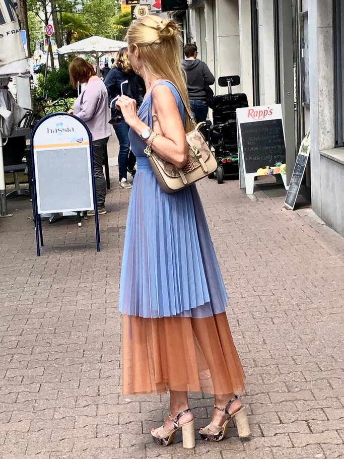 Blondhaarige Renate Zott im eleganten Plisseekleid in Blau und Rostbraun mit High Heels und Tasche spaziert über eine Einkaufsstraße. | © Renate Zott