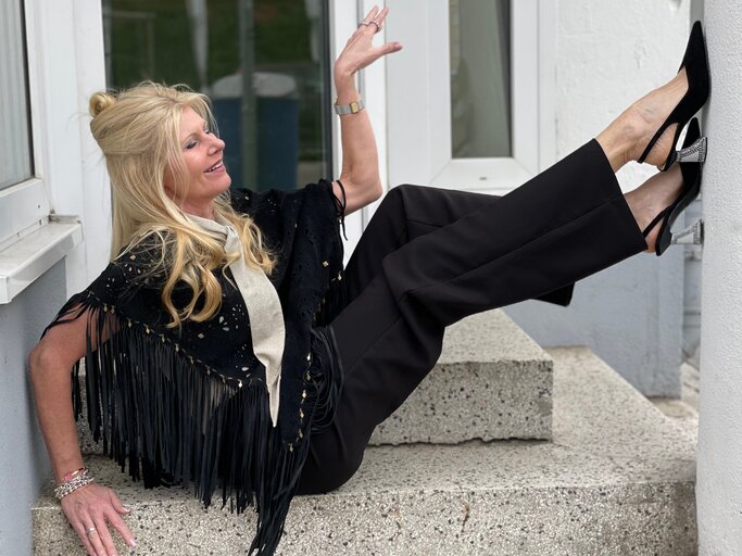 Blondhaarige Renate Zott in schwarzem Fransenponcho und Glitzerpumps lehnt lachend auf einer Steintreppe mit den Beinen in der Luft. | © Renate Zott