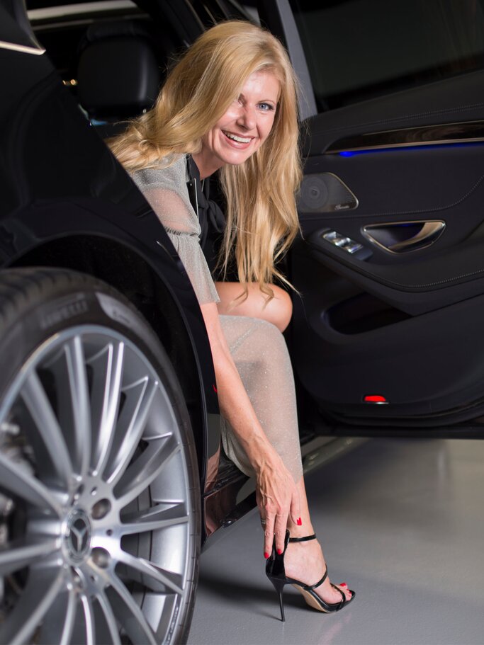 Blondhaarige Renate Zott im glitzernden Abendkleid steigt lächelnd aus einem schwarzen Auto und greift elegant an ihre High Heels. | © Sascha Scherer