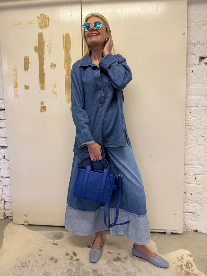Blondhaarige Renate Zott in langem Jeanskleid mit blauer Tasche und runder Sonnenbrille lächelt vor heller, grob gestrichener Wand. | © Renate Zott