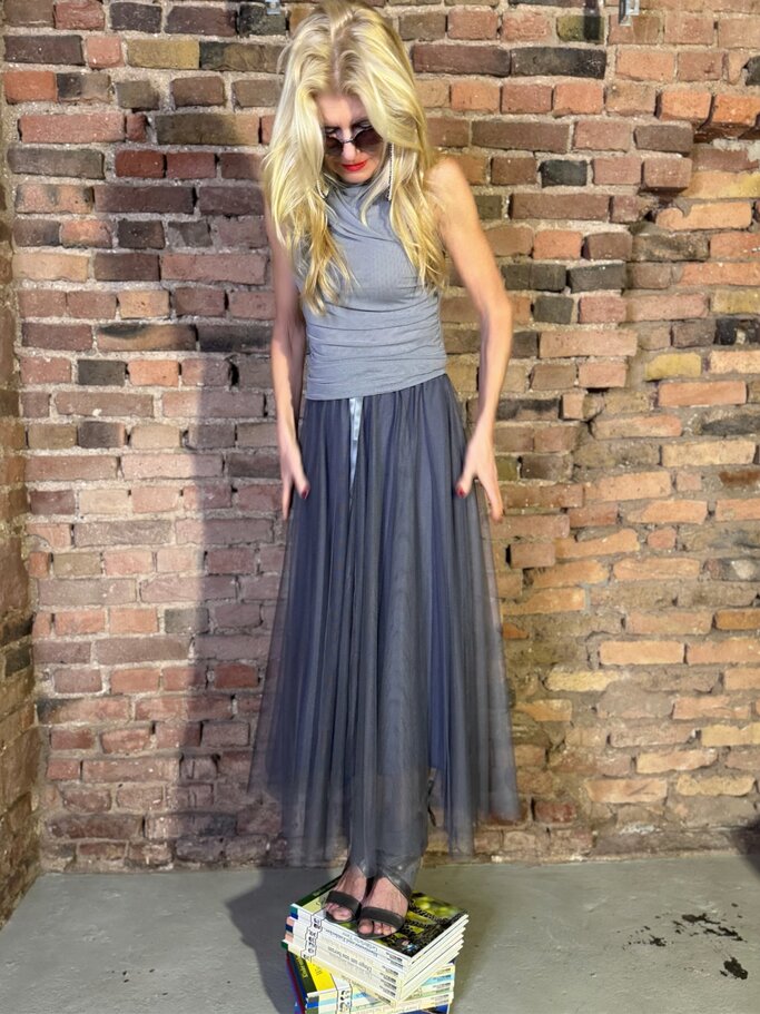 Blonde Renate Zott im ärmellosen Top, grauem Maxirock und schwarzen Sandalen balanciert auf Magazinen vor Backsteinwand. | © Renate Zott