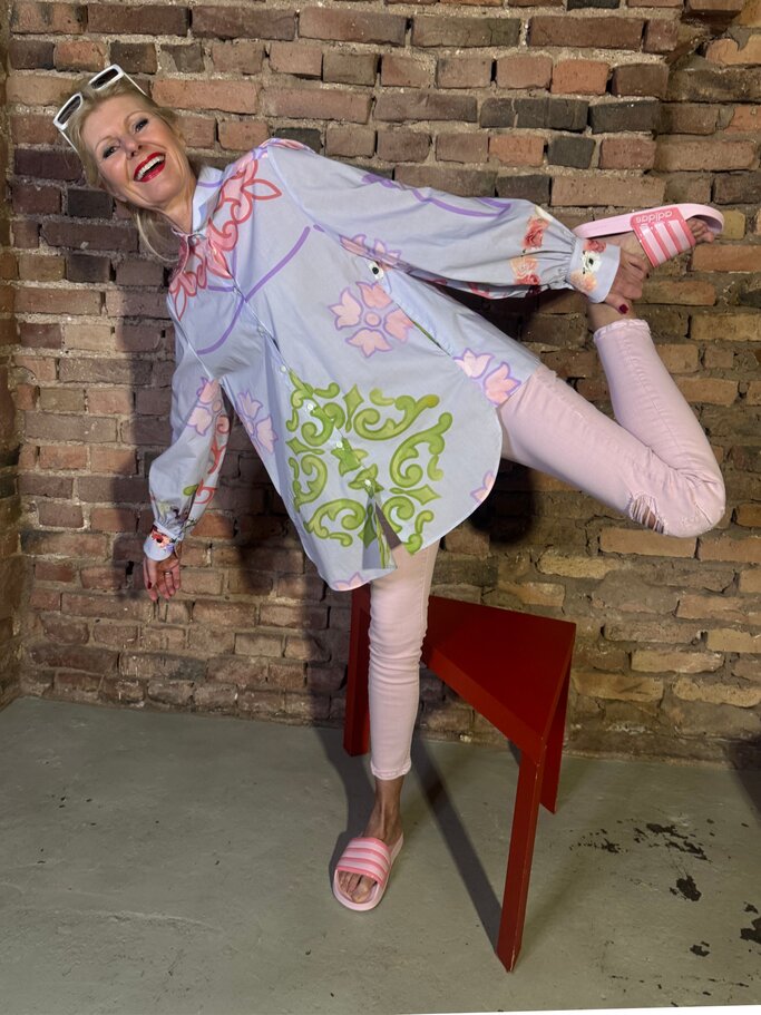 Renate Zott in pastellfarbenem Outfit mit floralem Muster steht lachend und locker vor rustikaler Ziegelmauer. | © Renate Zott Frühlings