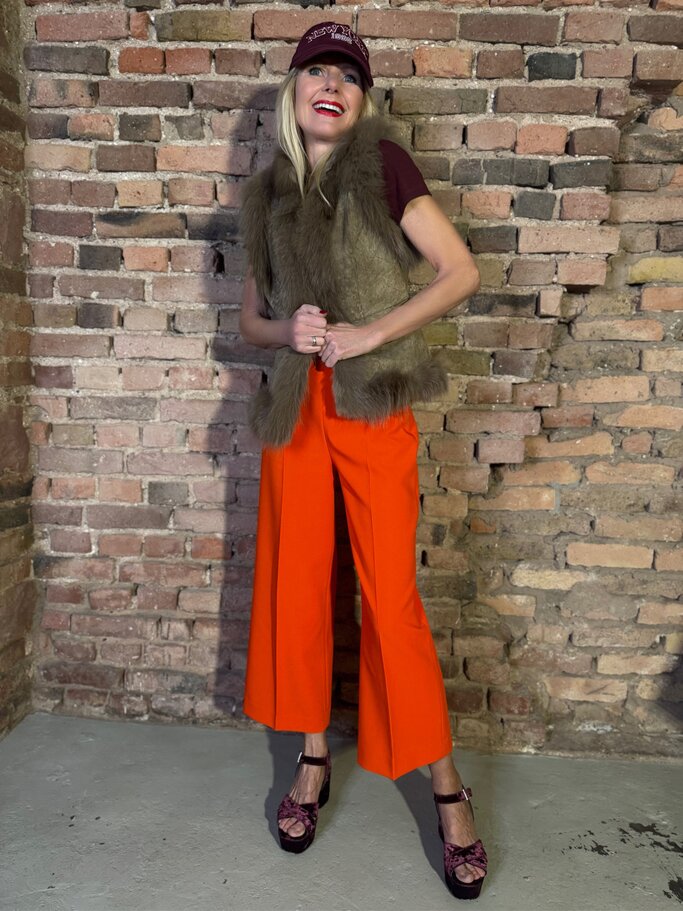Lachende Renate Zott mit bordeauxfarbener Kappe, Fellweste und orangefarbener Hose steht vor Backsteinwand. | © Renate Zott